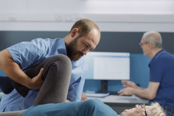 Medical specialist helping elder patient to cure physical injury with bones cracking procedure, stretching legs muscles in office. Therapist using alternative medicine to increase mobility.